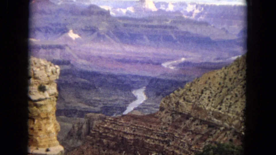 1967 8mm frame of the Grand Canyon in Arizona, showing a winding river through the canyon, rocky foreground walls, distant stratified formations, cloudy light, and dark rounded film borders.