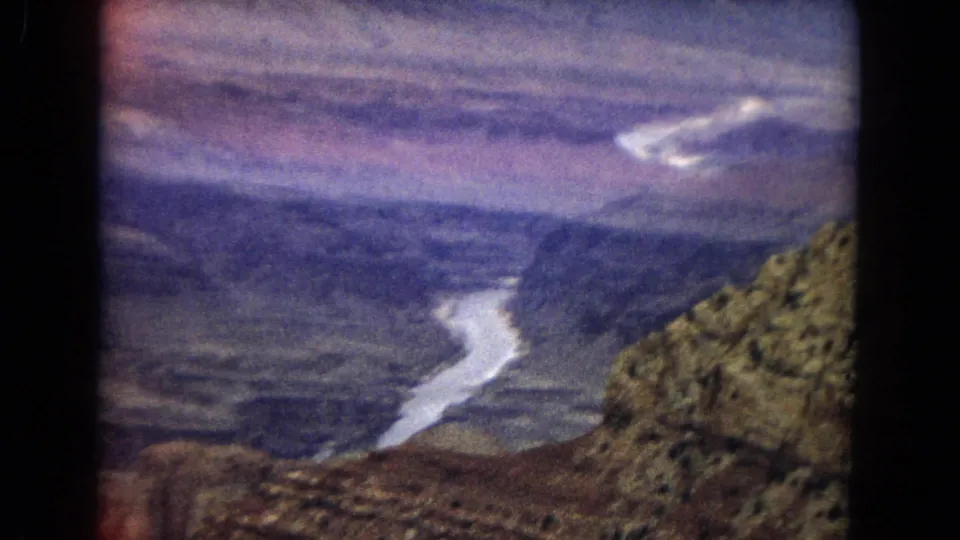 Handheld 1967 8mm view across the Grand Canyon, showing the Colorado River cutting through the canyon floor with layered rock formations and a scrubby slope in the foreground.
