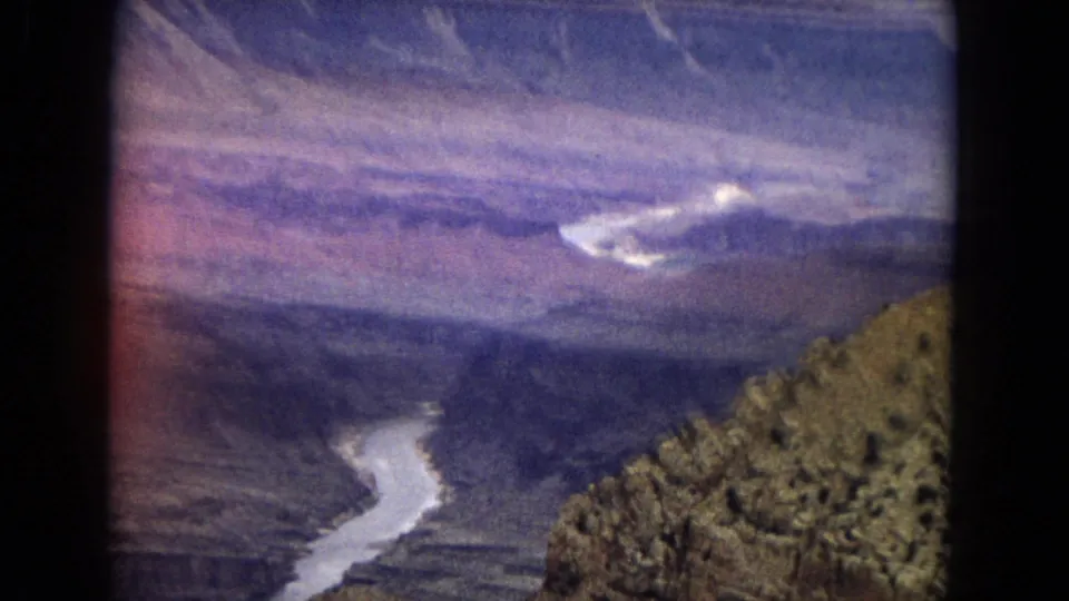 Vintage 8mm frame showing a soft, slightly hazy Grand Canyon panorama with the Colorado River below and a rocky, scrub-covered slope along the right foreground.