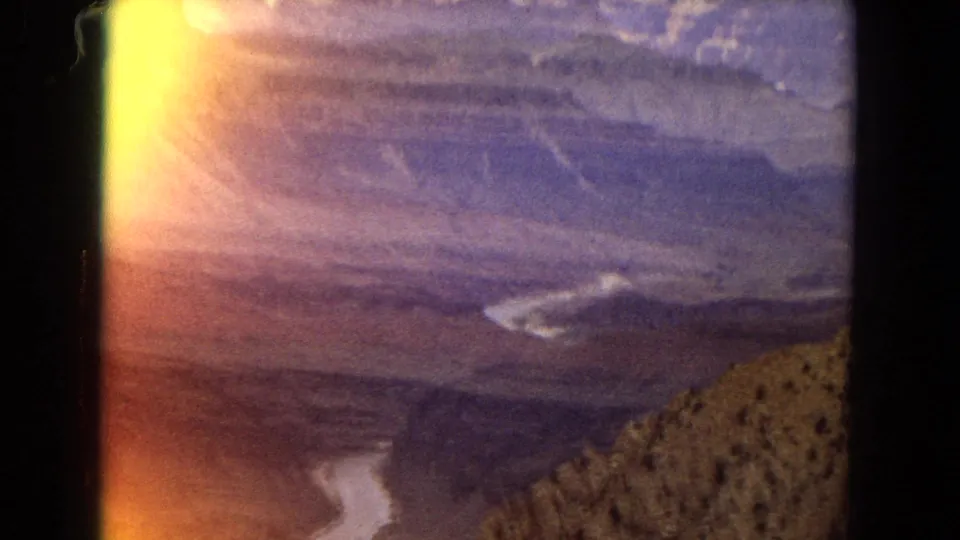 1967 8mm Grand Canyon view with the Colorado River below, layered cliffs in the distance, and a bright yellow-orange exposure flare along the left side of the frame.
