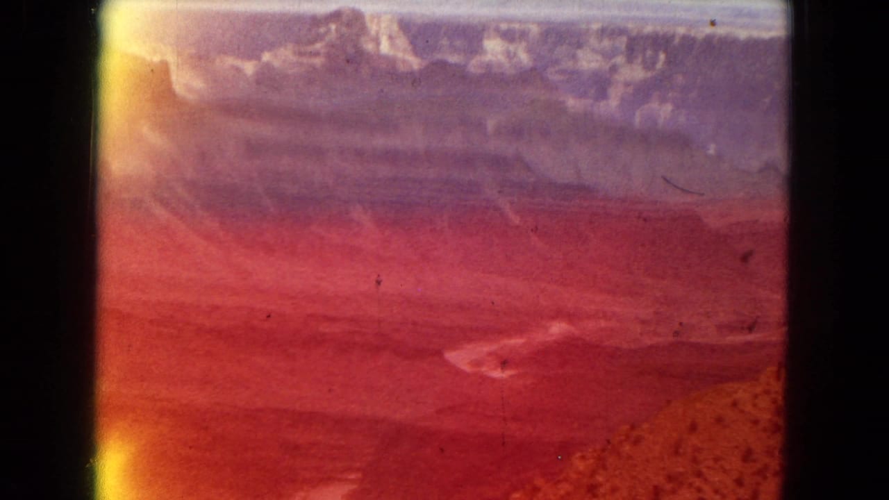 1967 8mm home-movie frame of the Grand Canyon area with a broad layered canyon view partly obscured by red-orange light leak and dark film borders.
