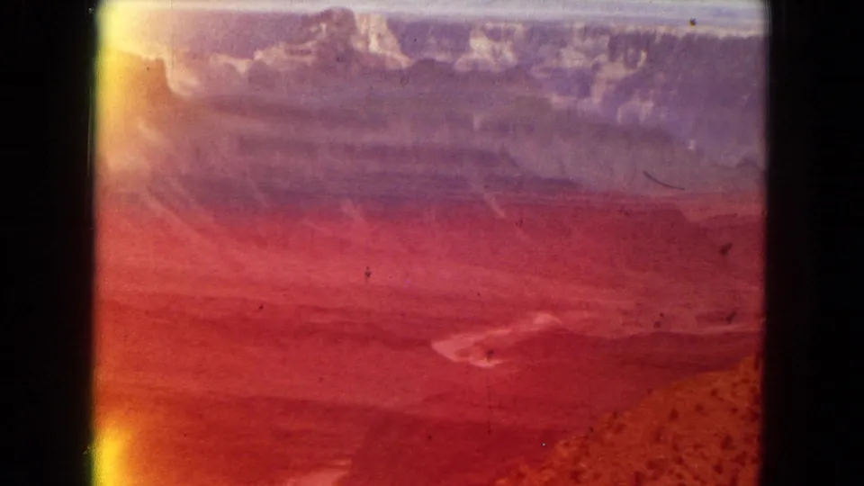 1967 8mm home-movie frame of the Grand Canyon area with a broad layered canyon view partly obscured by red-orange light leak and dark film borders.