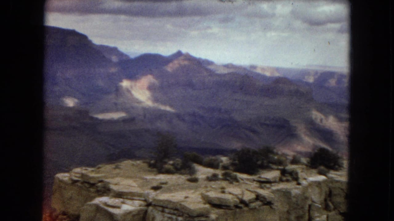 1967 Arizona travel frame showing a rocky Grand Canyon overlook with shrubs in the foreground and blue-gray canyon ridges receding under cloud cover.