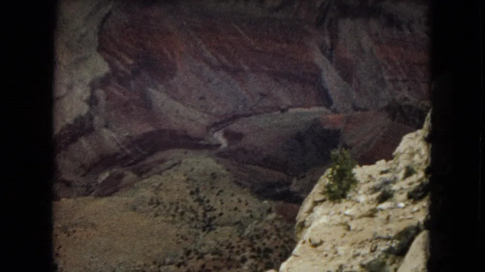1967 8mm view from a Grand Canyon rim ledge, looking across dark layered rock walls, a sloping canyon floor, and scattered desert vegetation.