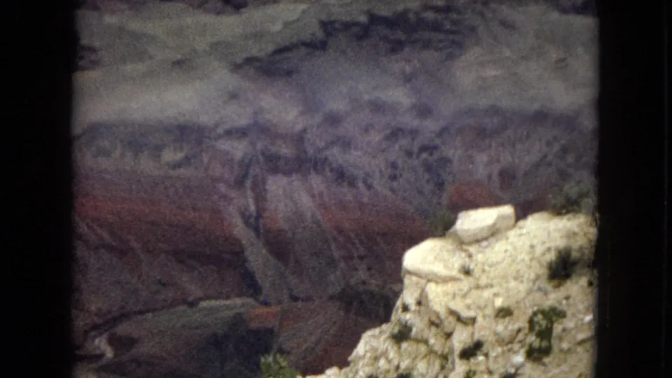 1967 Arizona home-movie frame of the Grand Canyon with a bright foreground rock outcrop, layered canyon walls, sparse shrubs, and clouds over distant cliffs.