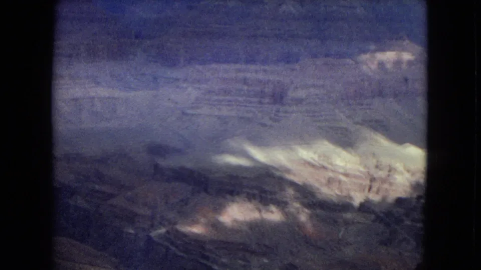 1967 8mm home-movie frame of the Grand Canyon in Arizona, with a pale ridge-like rock formation, broad shadowed canyon layers, and soft grain from the scanned film.