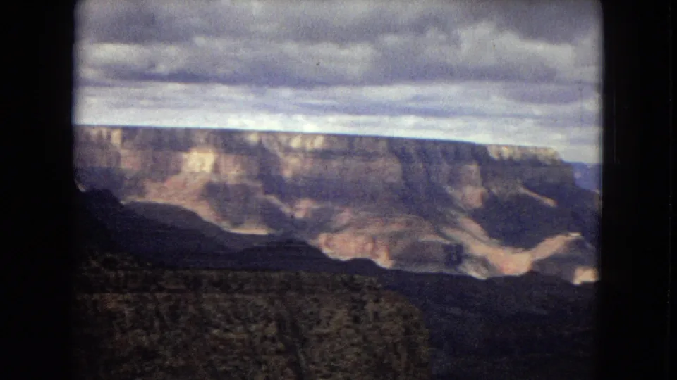 1967 8mm home-movie view of the Grand Canyon with layered cliffs, dark foreground terrain, and a dense overcast sky.