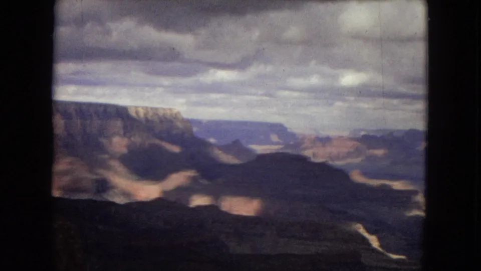 1967 8mm home-movie frame of the Grand Canyon with a shadowed valley, distant mesas, and soft cloudy light over the rock formations.