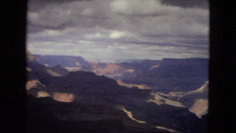 1967 8mm Grand Canyon view with dark ridgelines in the foreground, distant cliff bands, and gray clouds spanning the upper frame.