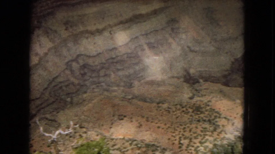 1967 Arizona 8mm frame showing a beige and gray striated rock wall with sparse scrub near the lower edge and dark cave-like borders from the film frame.