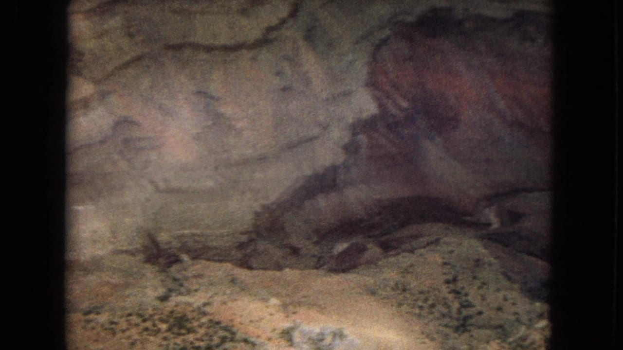 1967 Arizona home-movie frame of a shadowed cave or canyon wall with reddish and beige layered rock bands and a dark recess across the middle.