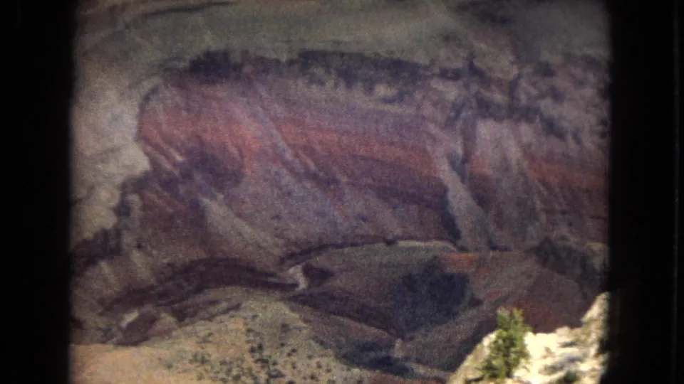 1967 Arizona 8mm frame showing reddish, purplish, and beige rock strata with a bright foreground ledge and a small patch of vegetation near the bottom edge.