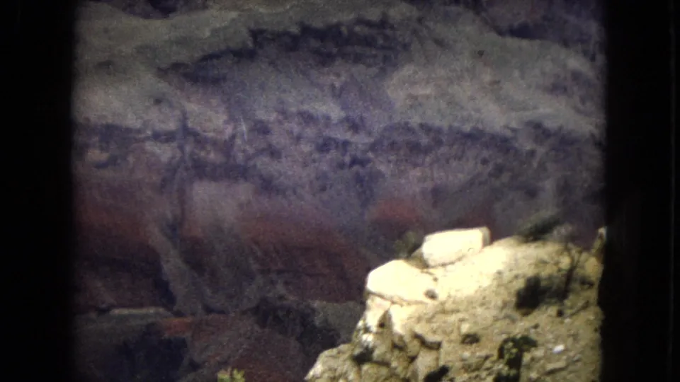 1967 Arizona home-movie frame with a pale sunlit boulder ledge in the foreground and a shadowed cave wall behind it marked by dark horizontal striations.