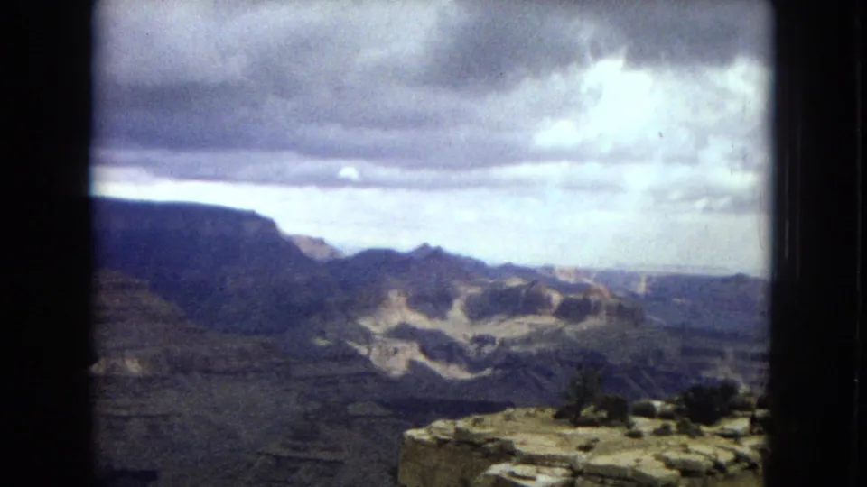 1967 Arizona home-movie frame looking across the Grand Canyon toward distant mesas beneath a heavy cloudy sky, with a rocky overlook edge in front.