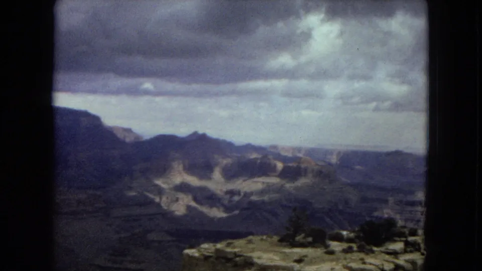 1967 Arizona 8mm frame of a broad Grand Canyon view with dark ridges, distant formations, low shrubs, and a rocky ledge beneath heavy clouds.