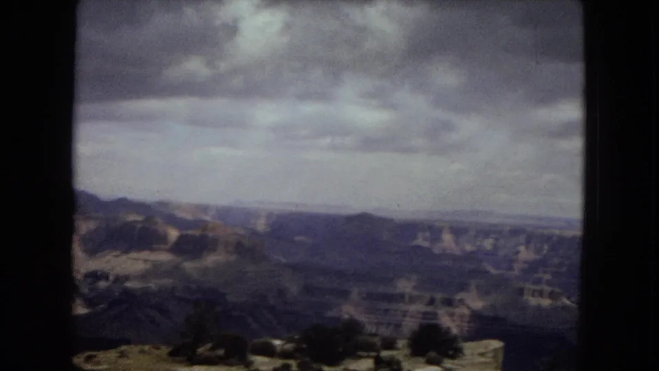 1967 Arizona home-movie frame showing a sweeping Grand Canyon horizon, layered canyon walls, sparse overlook vegetation, and a cloudy sky.