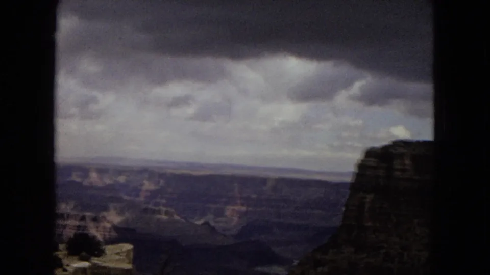 1967 Arizona 8mm frame showing the Grand Canyon with a dark cliff mass at right, distant canyon layers, and low overcast clouds.