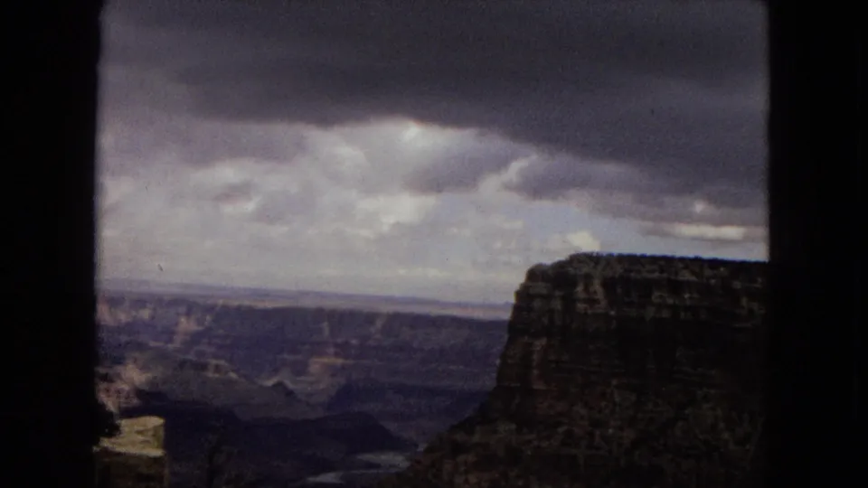 1967 Arizona 8mm frame with a moody Grand Canyon overlook, dark cloud cover, a foreground cliff, and layered ravines stretching into the distance.