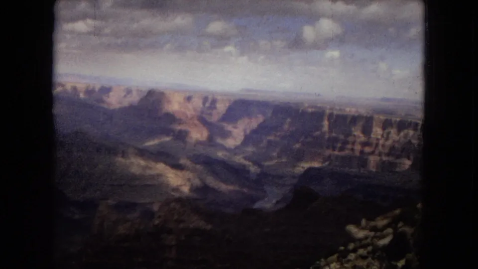 1967 Arizona 8mm frame showing layered Grand Canyon walls with partial sunlight, deep shadowed foreground rocks, and a pale clouded sky.