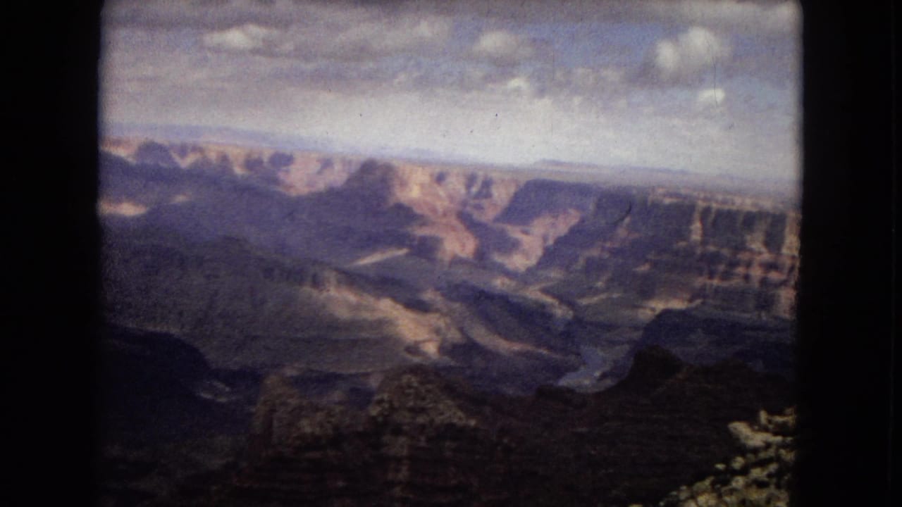 1967 Arizona 8mm frame showing a broad canyon view with layered rock walls, distant plateau forms, and a dark foreground overlook beneath overcast clouds.