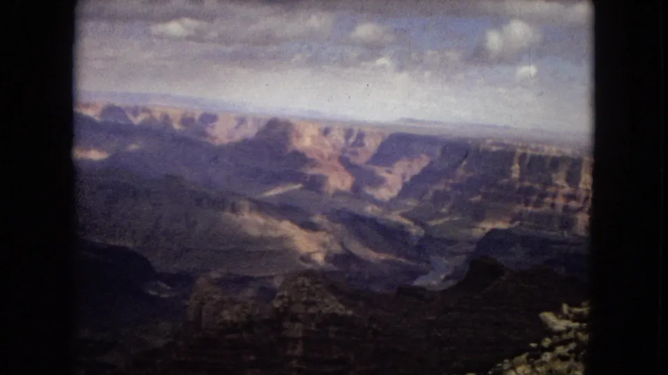 1967 Arizona 8mm frame showing a broad canyon view with layered rock walls, distant plateau forms, and a dark foreground overlook beneath overcast clouds.