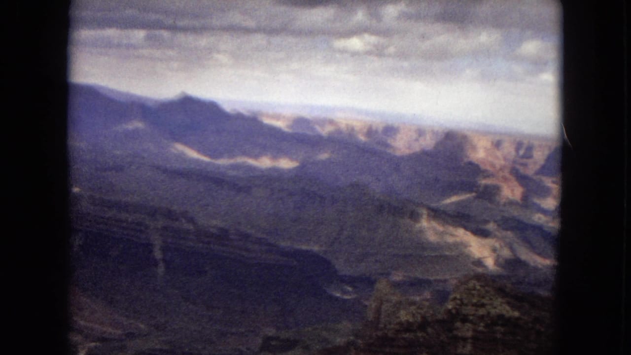 1967 Arizona 8mm frame with distant canyon ridges, shadowed rock formations, pale exposed slopes, and a cloudy sky over a remote landscape.