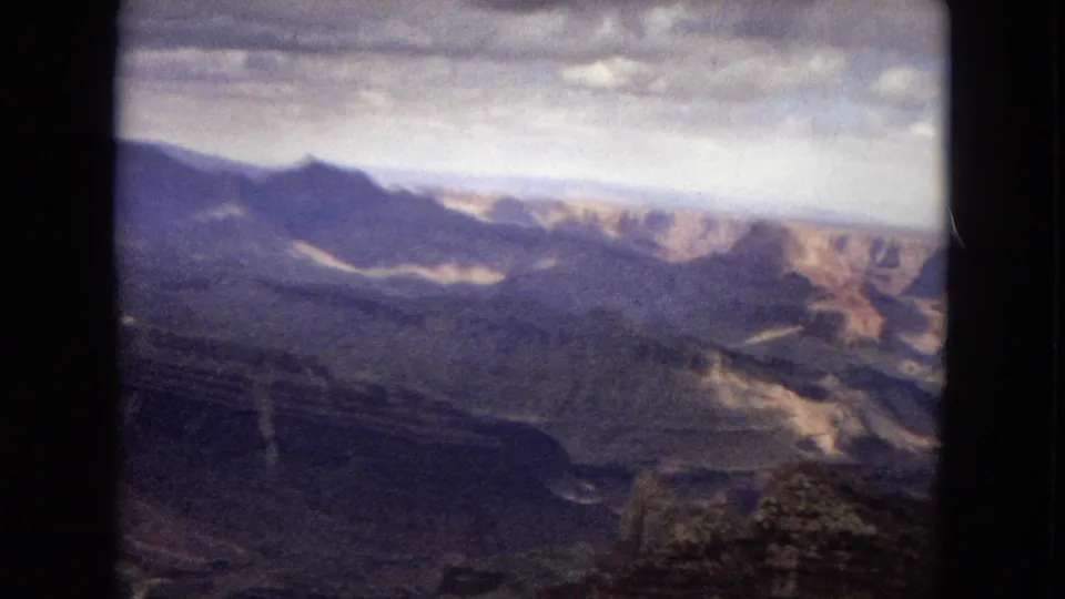 1967 Arizona 8mm frame with distant canyon ridges, shadowed rock formations, pale exposed slopes, and a cloudy sky over a remote landscape.