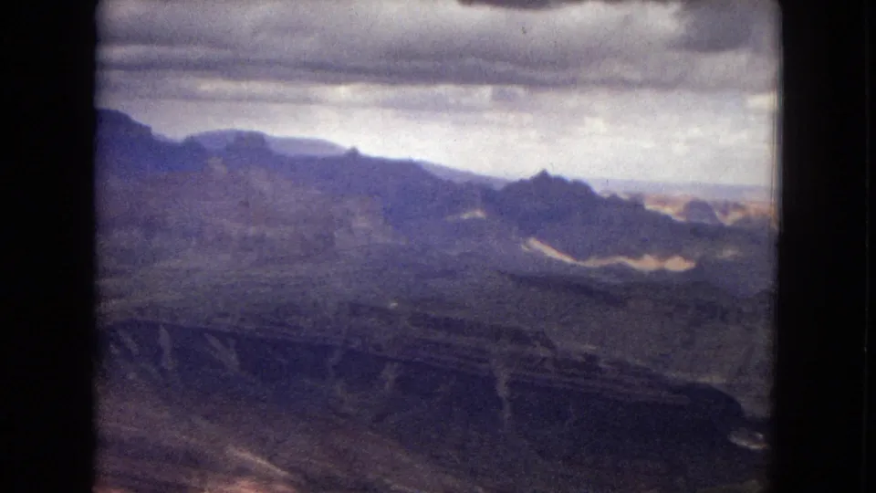 1967 Arizona 8mm frame showing an expansive canyon landscape with dark mesa shapes, layered slopes, a low horizon, and gray cloud cover.