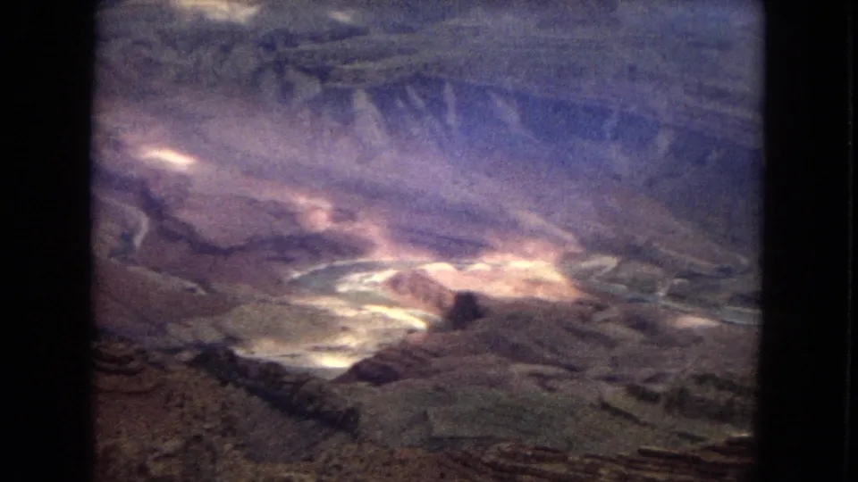 1967 8mm home-movie frame of an Arizona canyon view, with layered rock formations, a pale river course in the distance, cloudy sky, and dark rounded film borders.