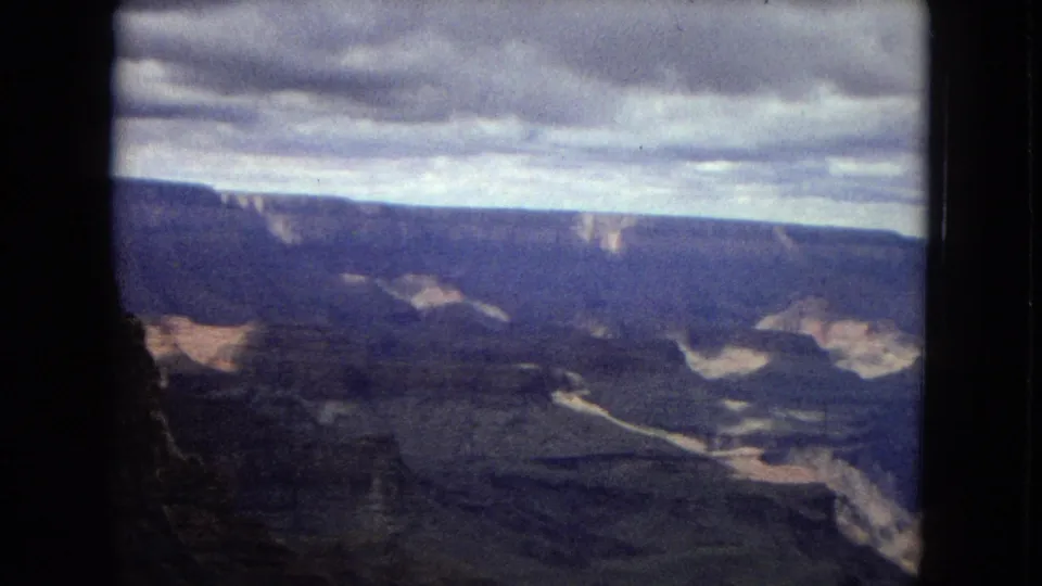 1967 8mm home-movie frame looking across the Grand Canyon area, with shadowed canyon depths, layered walls, a river trace far below, and a cloudy blue-gray sky.