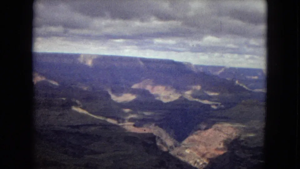 1967 8mm home-movie frame of an Arizona river canyon, showing a deep gorge, sunlit rock formations, distant mesas, heavy clouds, and soft vintage color.