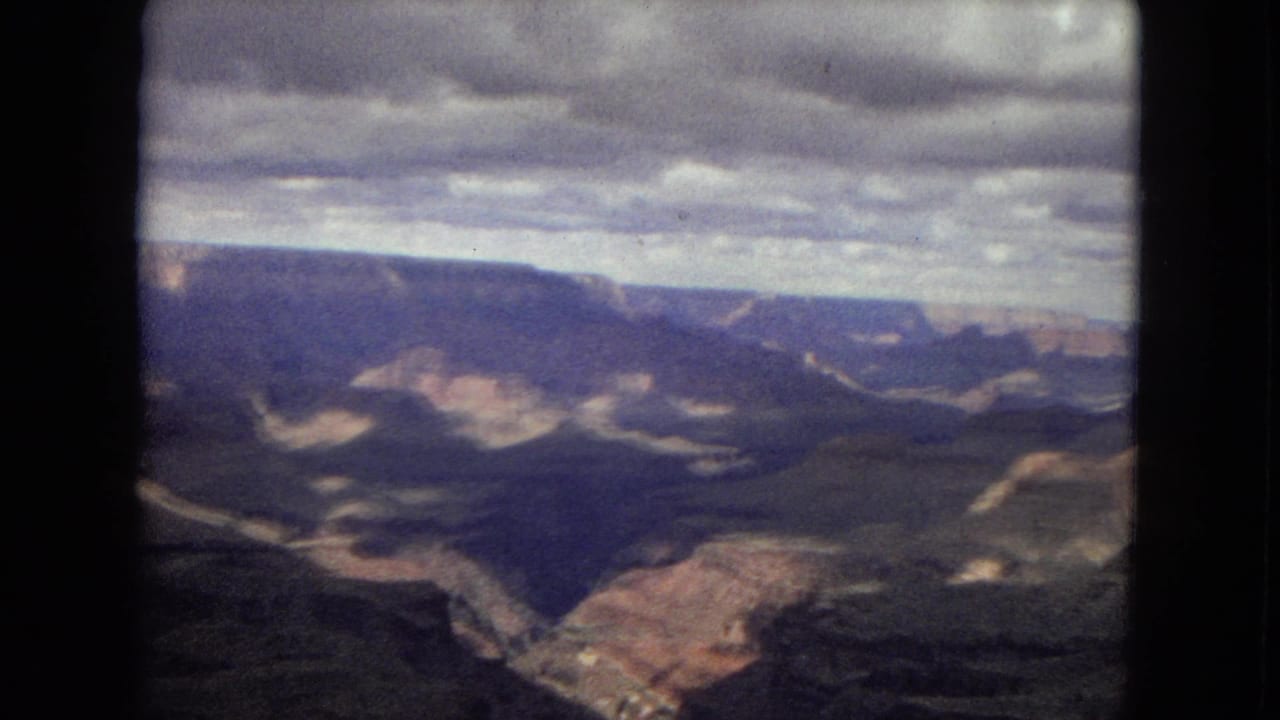 1967 8mm view of the Grand Canyon from an overlook, showing dark layered canyon walls, distant mesas, and a cloud-heavy sky.