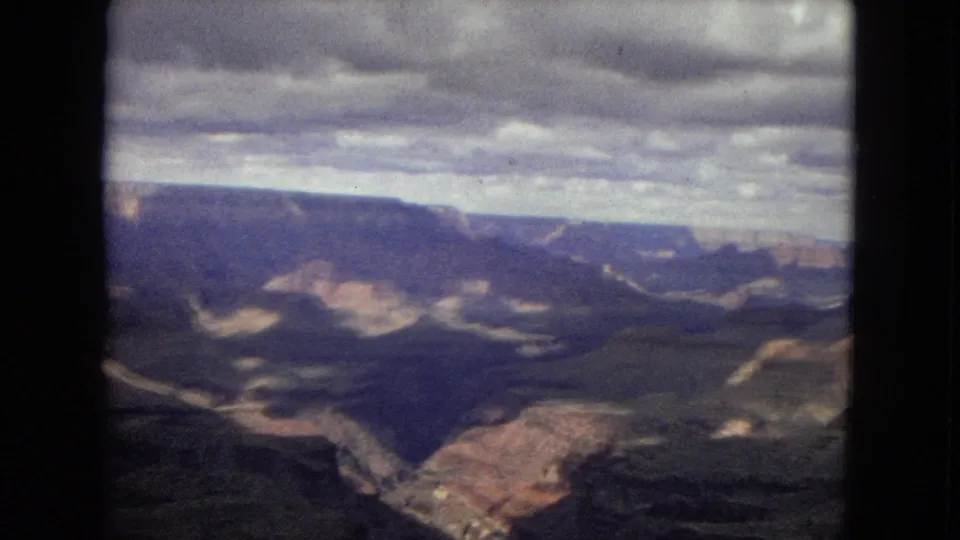 1967 8mm view of the Grand Canyon from an overlook, showing dark layered canyon walls, distant mesas, and a cloud-heavy sky.