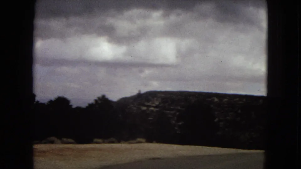 1967 Arizona travel footage frame showing a low desert hillside, tree silhouettes, and a heavy cloudy sky from a visitor viewpoint.