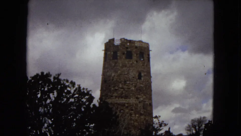 1967 Arizona home-movie frame showing a historic stone observation tower partially framed by trees beneath a cloudy sky.