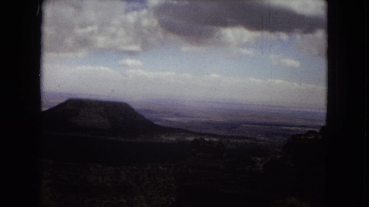 1967 Arizona travel footage frame showing a dark rounded hill or butte with distant plains and layered clouds beyond.