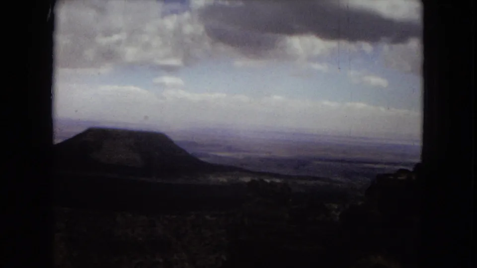 1967 Arizona travel footage frame showing a dark rounded hill or butte with distant plains and layered clouds beyond.