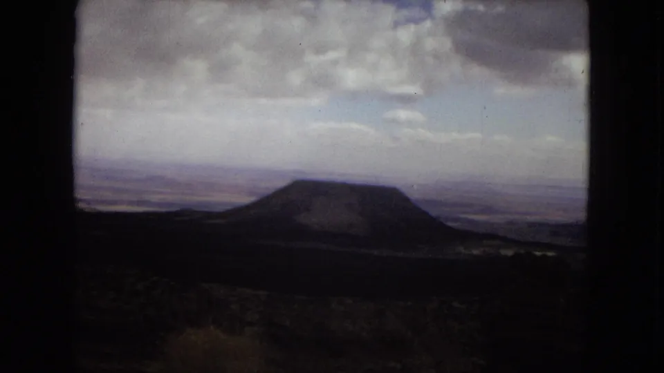 Wide 8mm home-movie view of an Arizona desert landscape with a rounded hill, dark foreground terrain, distant plains, and overcast sky.