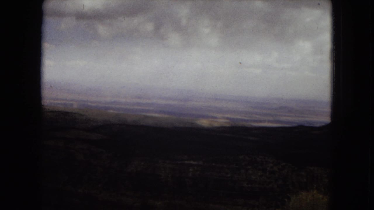 1967 8mm view in Arizona showing a shadowed canyon foreground, distant plateau horizon, and heavy cloud cover across the sky.