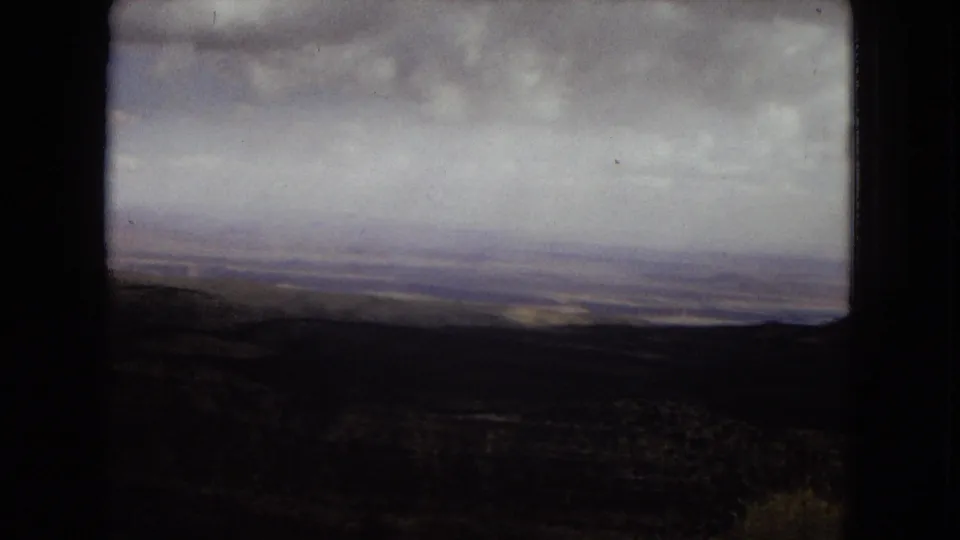 1967 8mm view in Arizona showing a shadowed canyon foreground, distant plateau horizon, and heavy cloud cover across the sky.