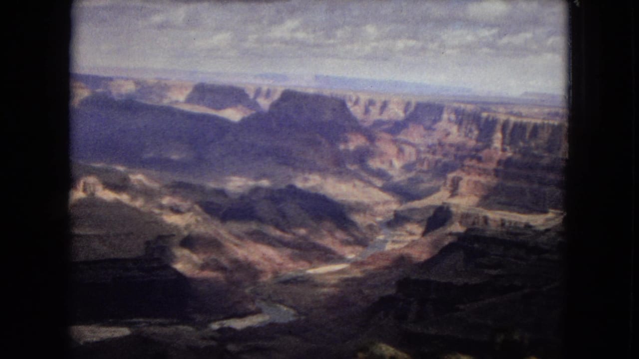 1967 8mm Grand Canyon view showing layered rock formations, shadowed ravines, and a broad overcast sky from a visitor viewpoint.