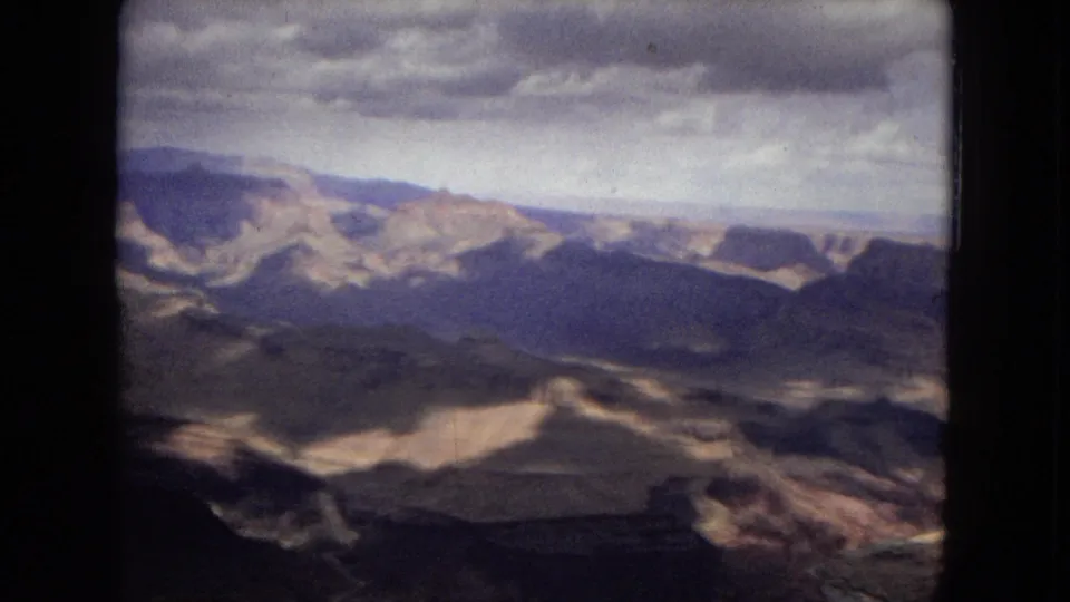 1967 8mm Grand Canyon frame showing dark foreground ridges, distant cliffs, layered rock color, and heavy clouds above the vista.
