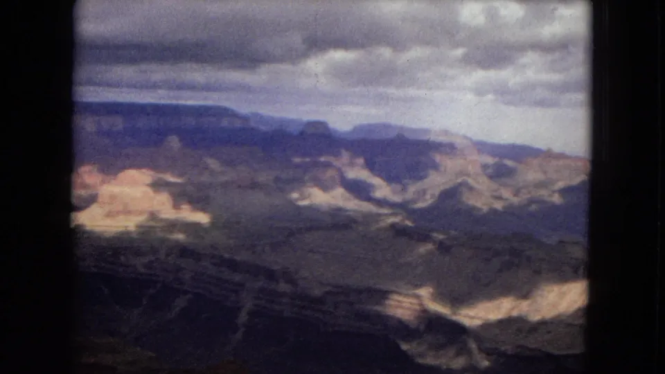 1967 8mm Arizona view across the Grand Canyon, with distant plateau edges, shadowed rock layers, and patches of pale sunlight.