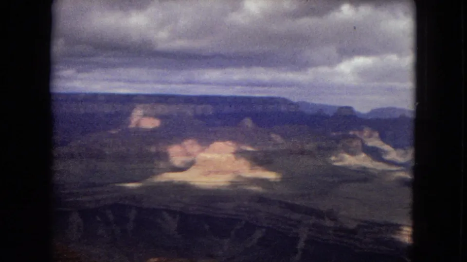 1967 8mm Arizona frame showing the Grand Canyon from an overlook, with horizontal rock layers, pale formations, and low gray clouds.
