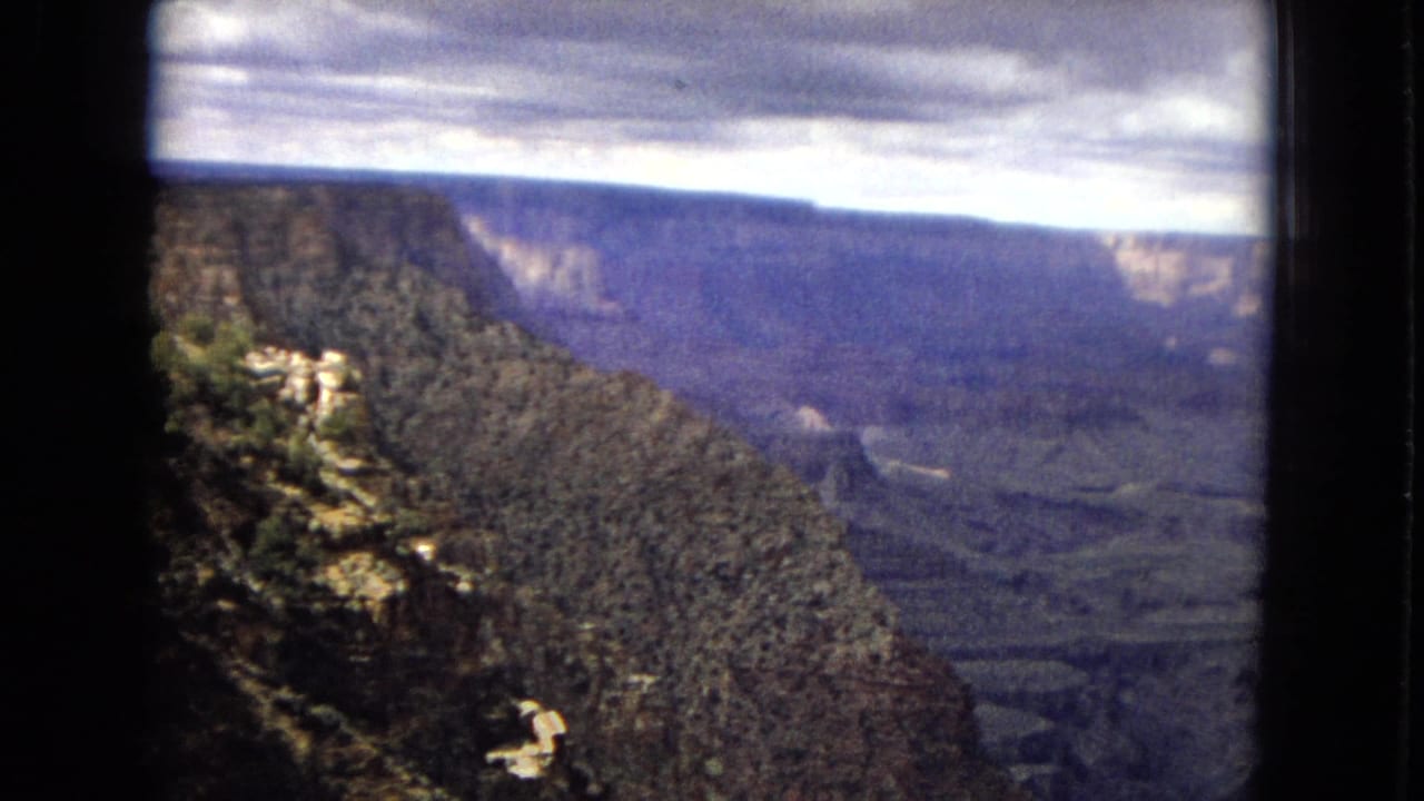 1967 8mm home-movie frame of a Grand Canyon overlook in Arizona, with a dark forested slope in the foreground and layered canyon walls under heavy clouds.