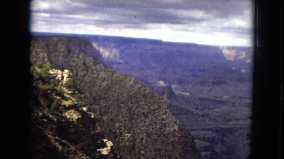 1967 8mm home-movie frame of a Grand Canyon overlook in Arizona, with a dark forested slope in the foreground and layered canyon walls under heavy clouds.