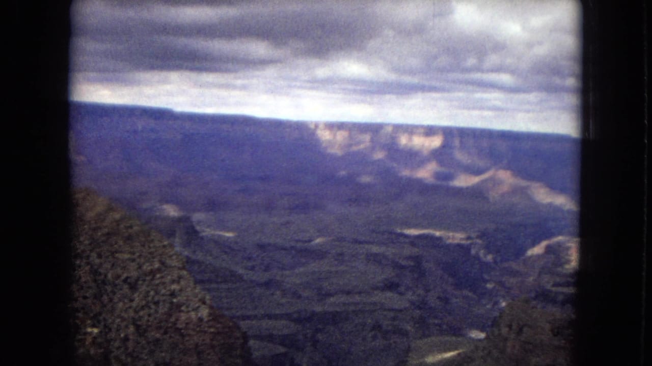 1967 8mm home-movie view across the Grand Canyon in Arizona, showing shadowed canyon terrain, pale cliff bands, and an overcast sky within visible film-frame borders.
