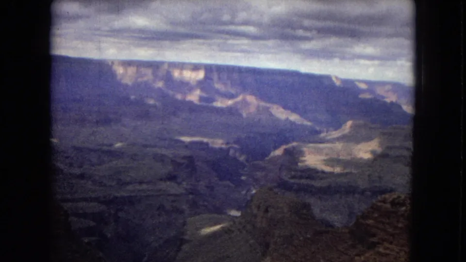 1967 Arizona Grand Canyon frame showing layered rock terraces, distant cliff faces, and a muted cloudy sky in soft, grainy 8mm color.