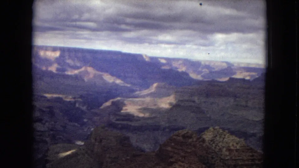 1967 8mm home-movie frame from Arizona showing the Grand Canyon with foreground rock outcrops, distant buttes, layered walls, and a cloudy horizon.