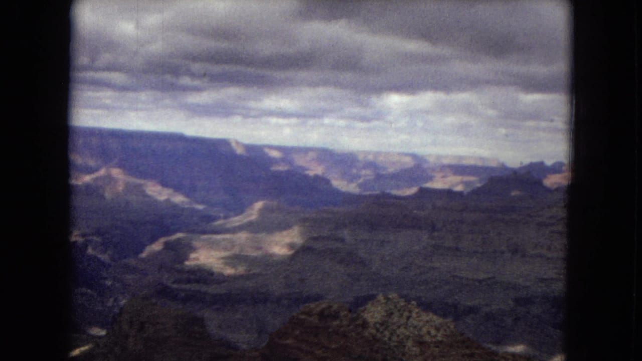 1967 Arizona 8mm frame showing a broad Grand Canyon vista with layered rock walls, a rocky foreground ridge, and dense overcast clouds above the horizon.